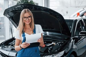 Manager with notepad, in the white shirt and blue uniform stands against automobile with hood up